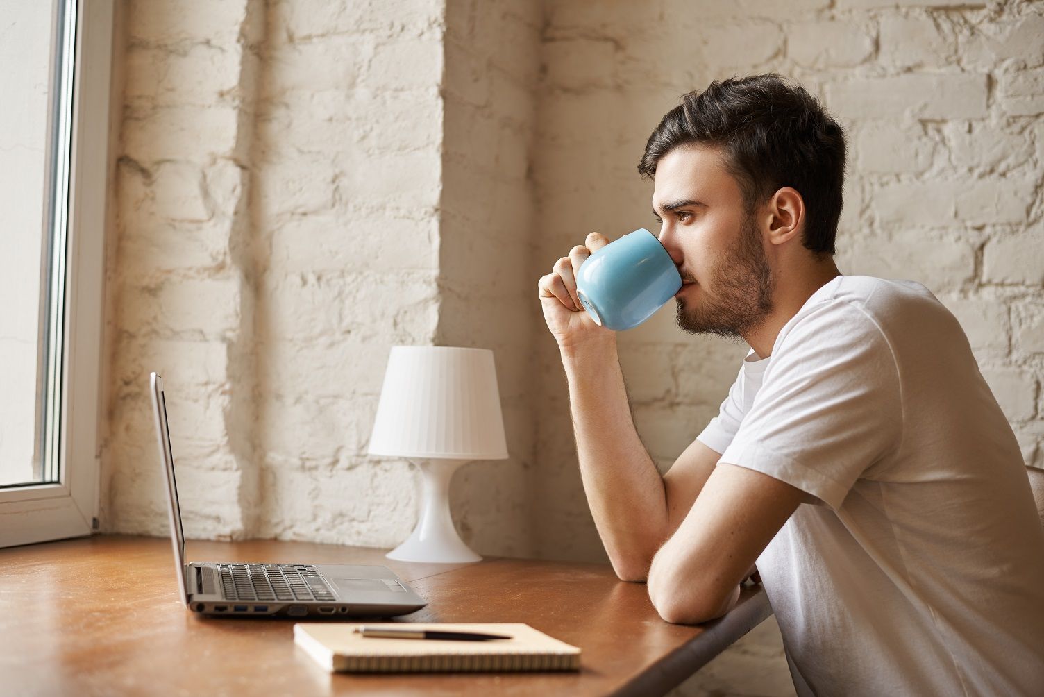 Handsome hipster drinking coffee after freelance work. Man in white t-shirt look to the window and thinking about new strategy to earn more money. Student search job in the internet.